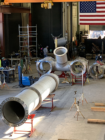 PMI's Sacramento pipe fabrication shop is seen from above. A large, custom fabricated pipe assembly sits in the middle of the shop, with pipe fabricators and an American flag in the background.